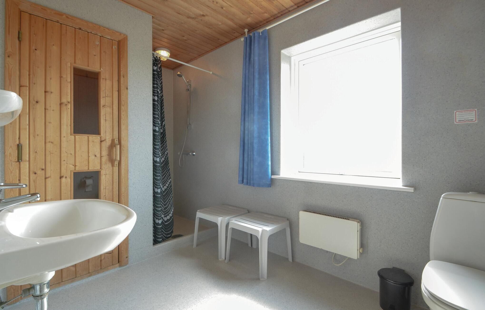 Un baño con puerta de madera, ducha, ventana con cortina azul, lavamanos, dos taburetes blancos y un radiador.