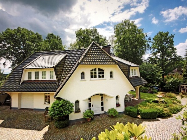 Une maison traditionnelle avec un toit à forte pente, de nombreuses fenêtres et un jardin bien entretenu, agrémenté de haies et d’une fontaine.
