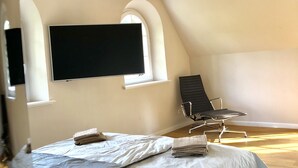 Ein Schlafzimmer mit einem großen Bett, einem Sessel, einem an der Wand befestigten Fernseher und einem Fenster.