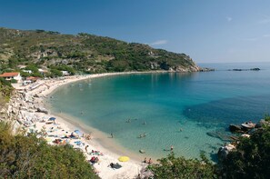 Ein Küstenstrand mit klarem türkisblauem Wasser, sandigem Ufer und hügeliger Kulisse.