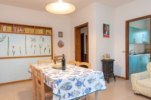 Un comedor con una mesa dispuesta para dos personas, una estantería, un cuadro y una cocina visible a través de un umbral abierto.