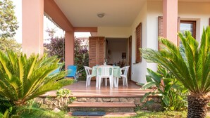 Eine überdachte Terrasse im Freien mit Tisch und Stühlen, umgeben von Grünpflanzen und einem gepflasterten Weg.