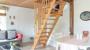 En trætrap fører op til en overetage, et spiseområde med et bord og stole, en sofa med puder, et lille bord med en vase og et billede i ramme på væggen.