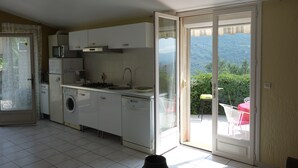A compact kitchen with basic appliances, a dining area with a table and chairs, and a sliding glass door leading to a balcony with a view of greenery.