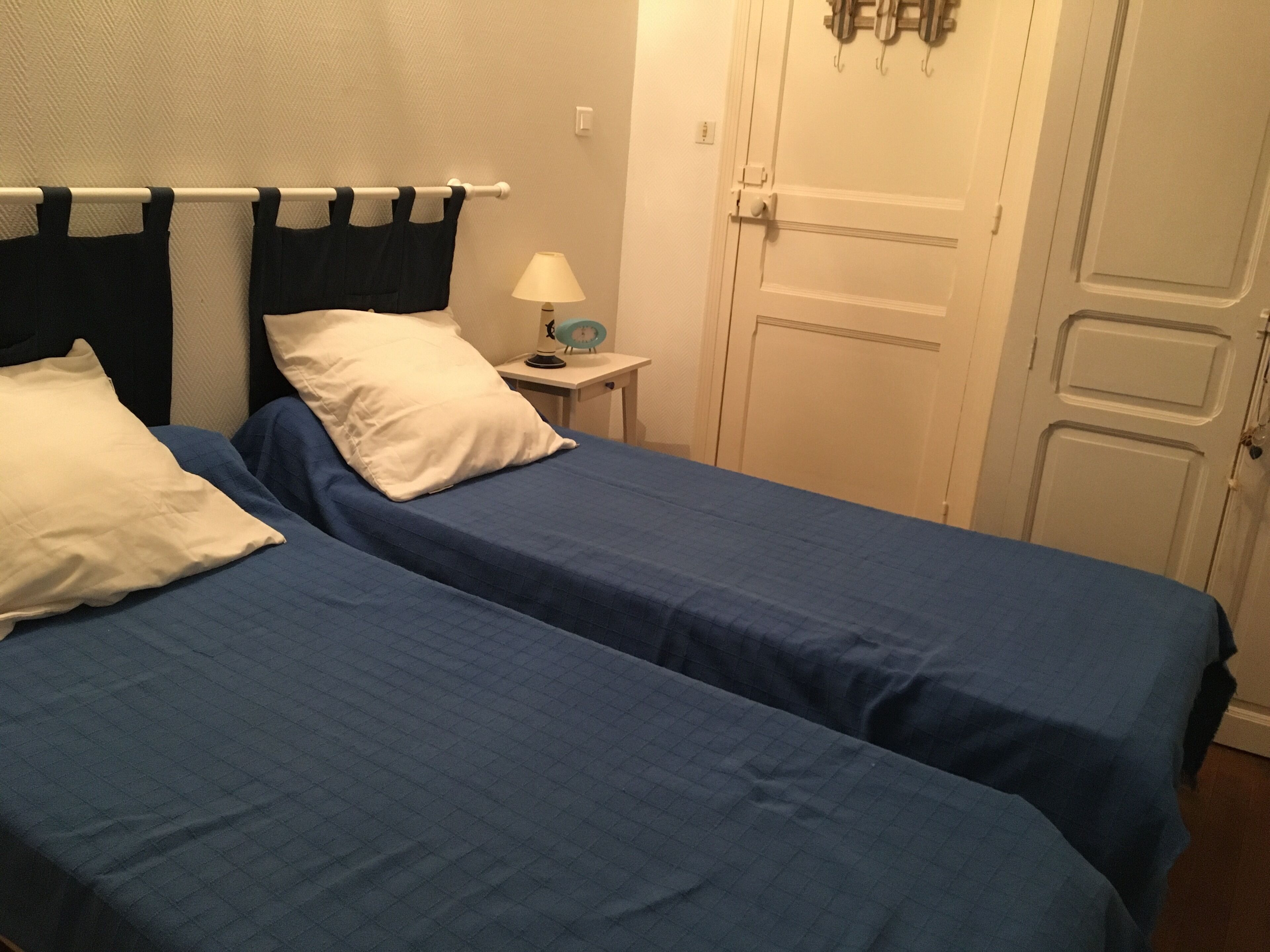 A room with two beds, each with a blue bedspread and white pillows. There is a small bedside table with a lamp and a clock. The room has white walls and a white door with a handle.