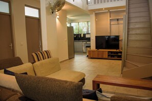 A living room with a sofa, a television on a wooden stand, and a kitchen in the background.