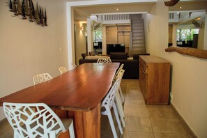 Un comedor con mesa de madera y sillas blancas, una zona de cocina con estufa y alacenas, y un área de estar con sofá y televisor.