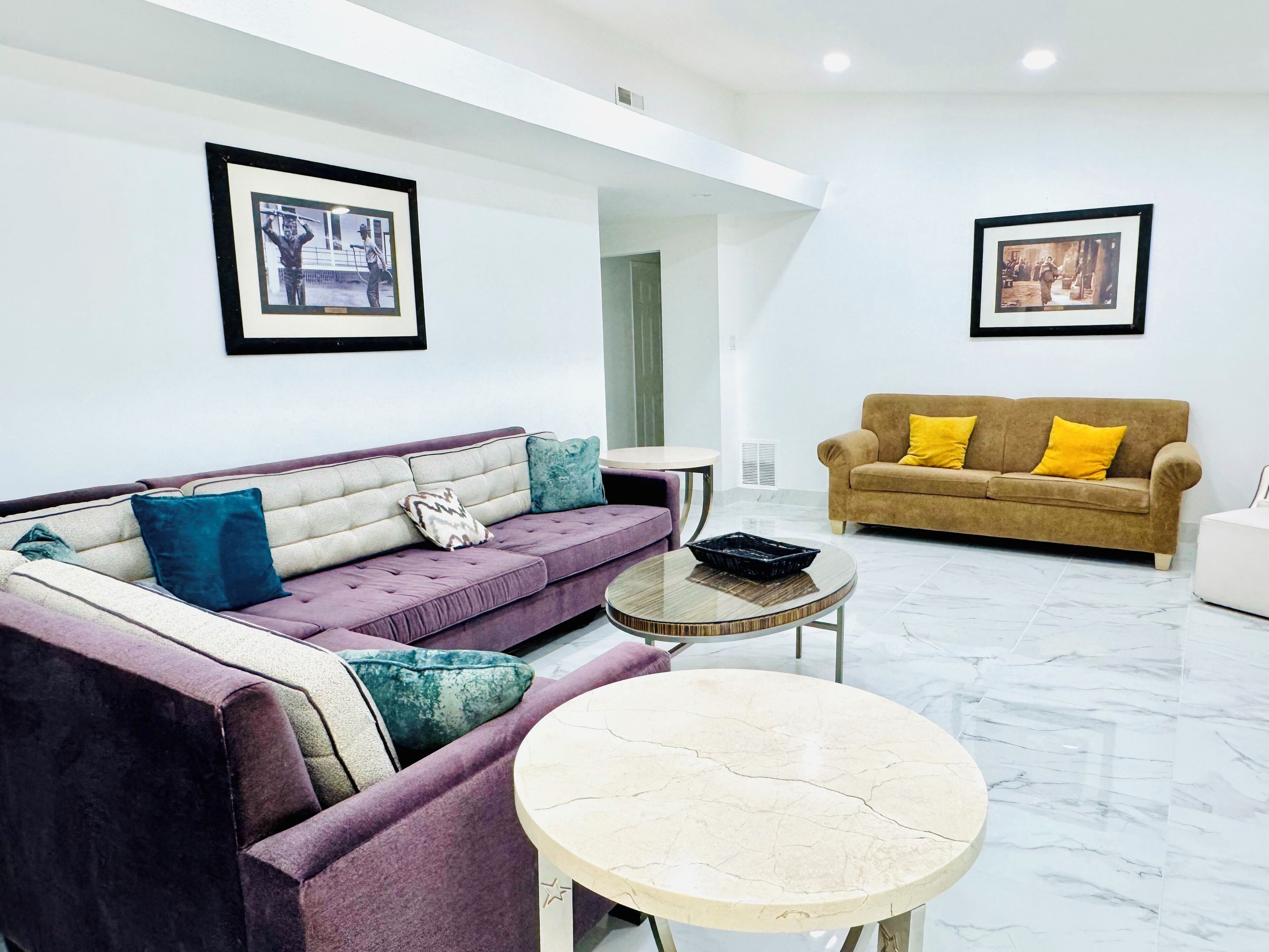 A modern living room with a purple sofa, a round coffee table, and a brown sofa with yellow cushions. There are framed pictures on the walls.