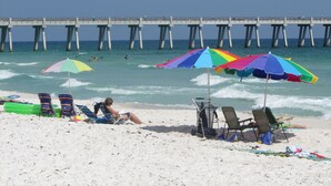 A beach with umbrellas, chairs, and a pier in the background. - The Pearl #304 - FREE BEACH SETUP March-October! (Navarre Beach)