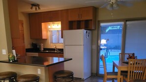 A kitchen with wooden cabinets, a white refrigerator, and a bar area with stools. There is a dining table with chairs and a view of a swimming pool through a sliding glass door. - Pool House - 3-bdrm, 2 bath, Heated Pool, Walk to Asu (Tempe)