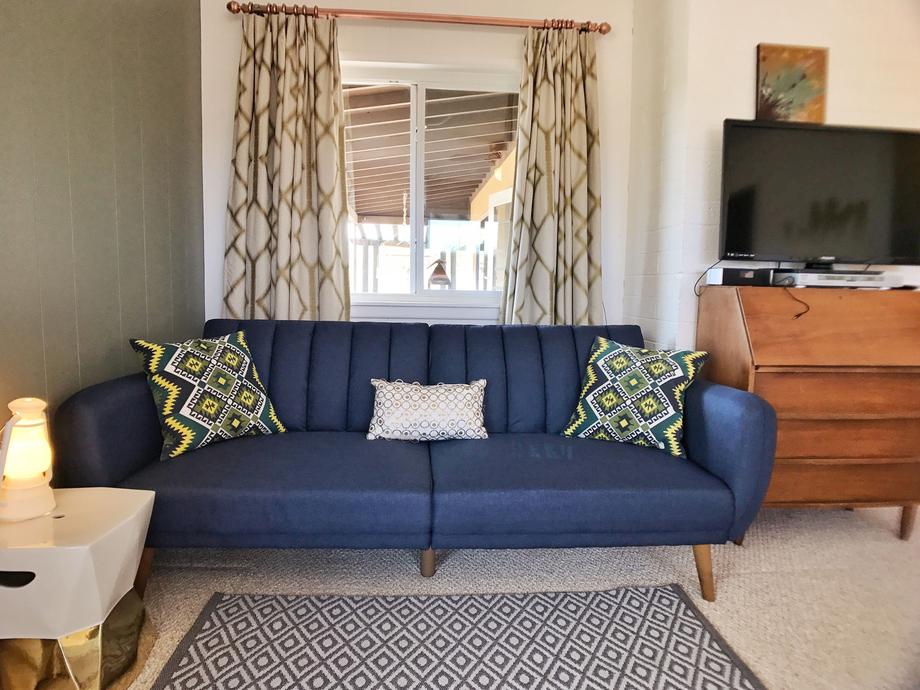 Un salon avec un canapé bleu foncé, des coussins à motifs, un tapis à motifs, une commode en bois et un téléviseur posé sur le dessus.