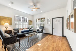 A living room with a leather sofa, a coffee table, and a black armchair. There is a ceiling fan, a window with blinds, and a door with a mirror.