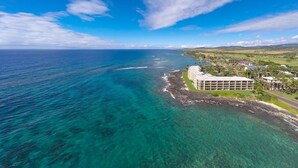 An aerial view of a coastal hotel with clear turquoise waters, a rocky shoreline, and a large building with multiple floors. - Live TOP Floor OceanFRONT. Relax & Sleep to Sounds of the Surf; AC or Tradewinds (Koloa)