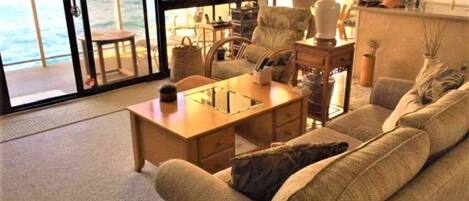 Un salón acogedor con sofá, mesa de centro y sillones, una lámpara y vista al océano a través de amplias ventanas.