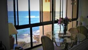 A room with a large window overlooking the ocean, a round table with chairs, and a vase with flowers. - Live TOP Floor OceanFRONT. Relax & Sleep to Sounds of the Surf; AC or Tradewinds (Koloa)