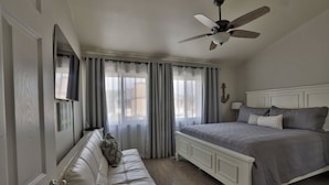 A bedroom with a bed, a white headboard, a ceiling fan, a window with curtains, a wooden floor, and a door with a handle. - Walk To Disneyland, Disneyland Rental Home 2. (Anaheim)