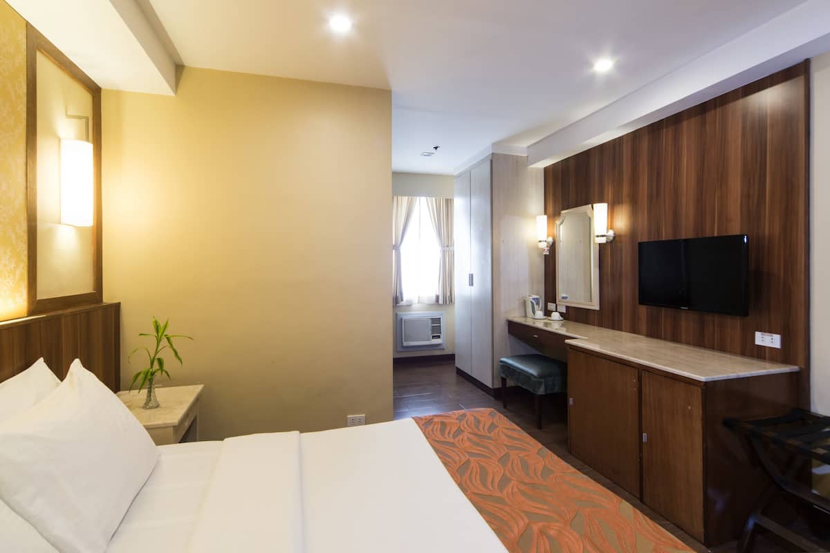A hotel room with a bed, a desk, a TV, and a bathroom visible in the background.
