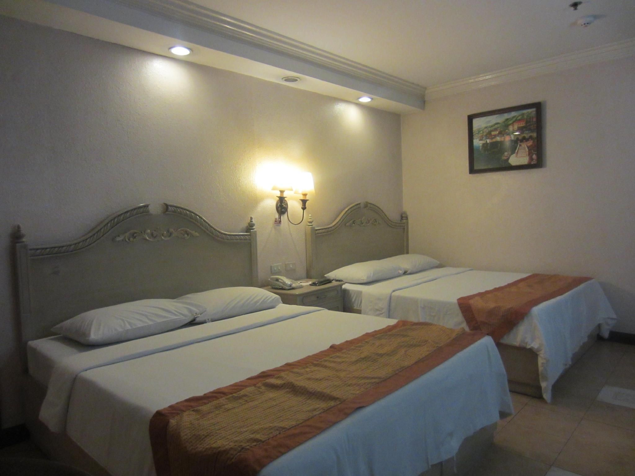 A hotel room with two single beds, a nightstand with a lamp, a painting on the wall, and a ceiling light.