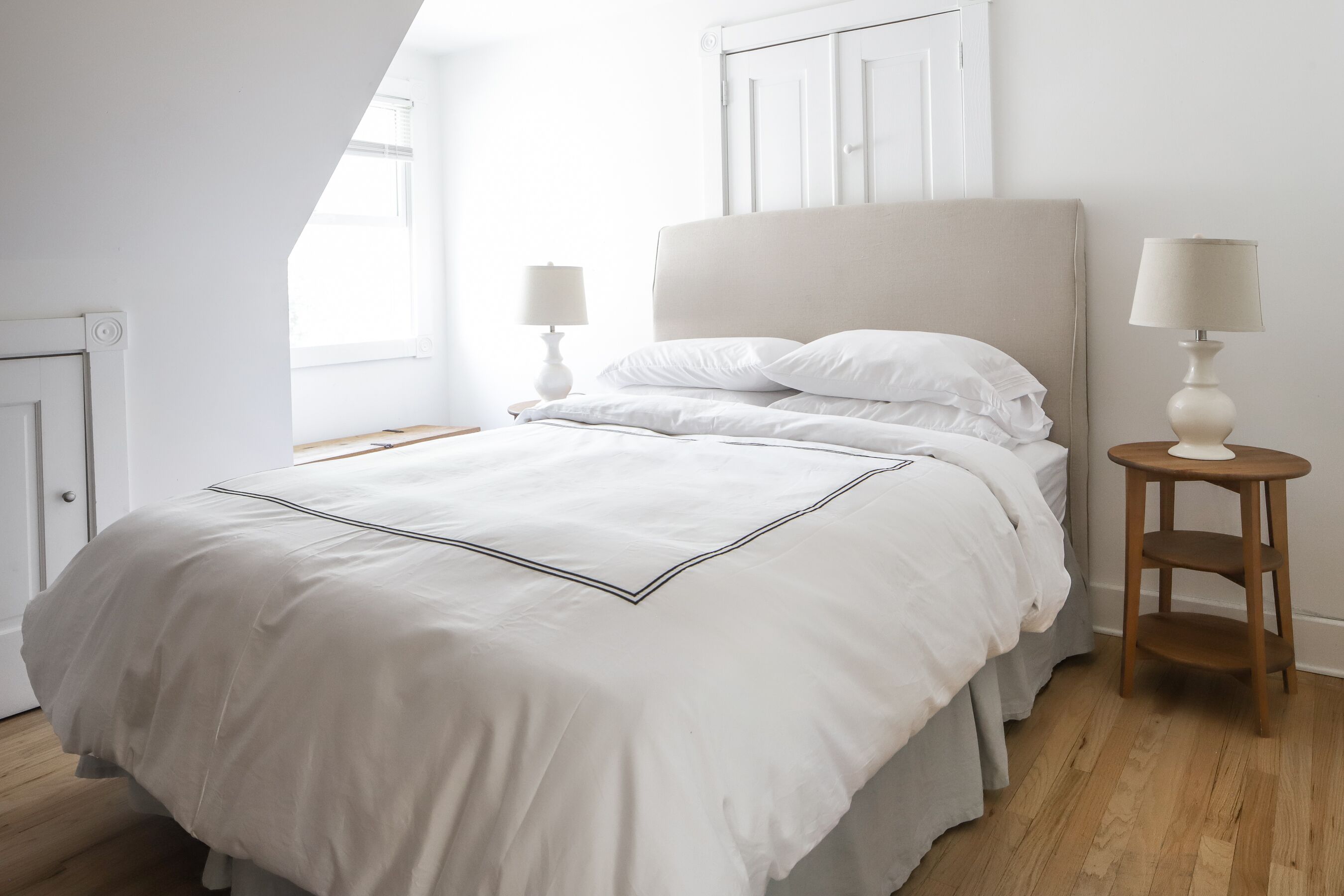 A neatly made bed with a white duvet and pillows, two bedside tables with lamps, and a window with blinds.