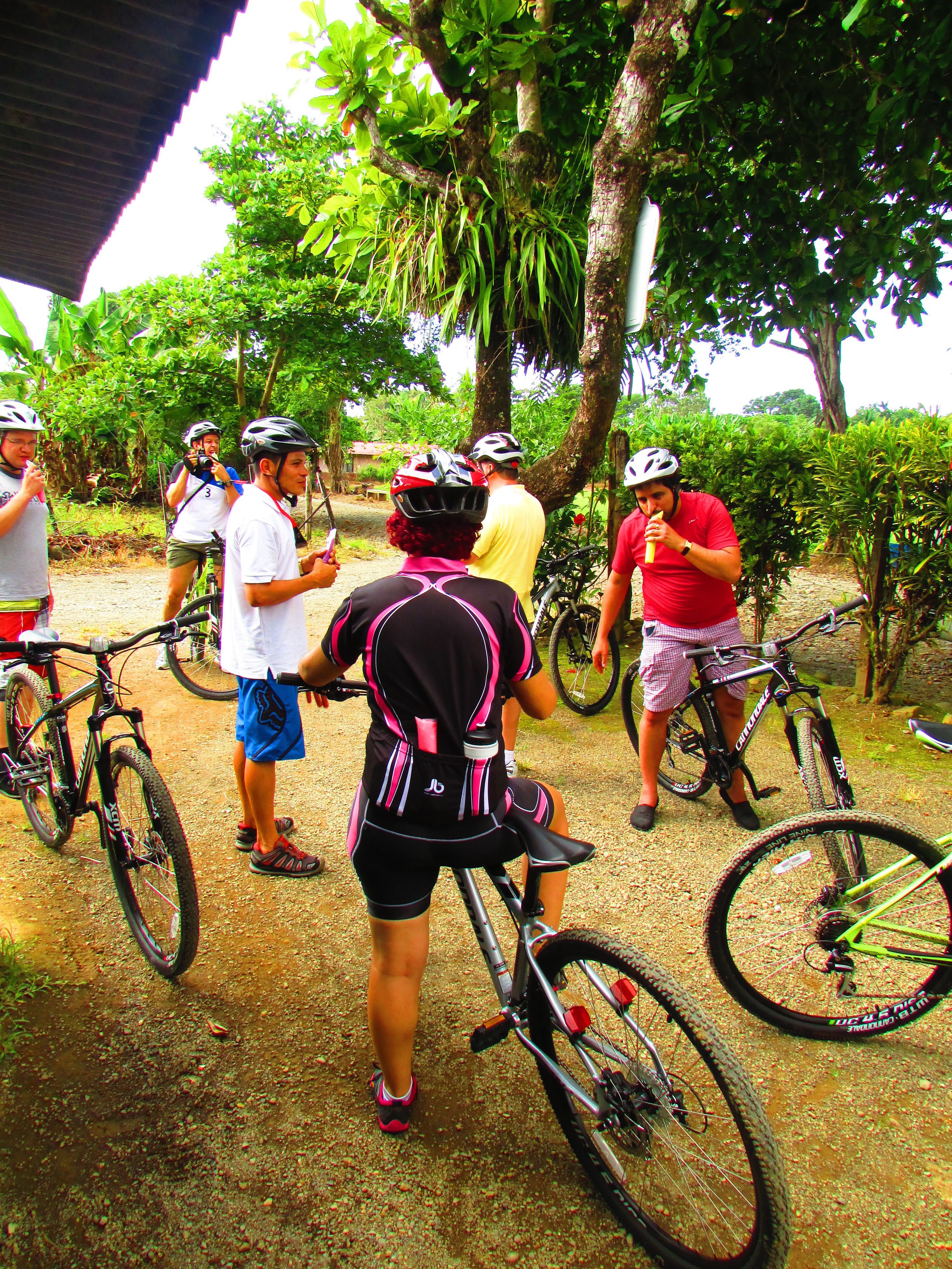 A group of people with bicycles in a shaded outdoor area with trees and a dirt ground.