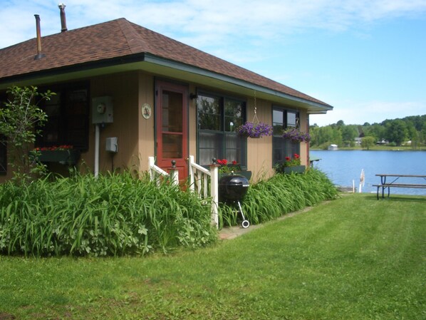 Un edificio junto al lago, con puerta roja, ventanas grandes y macetas colgantes de flores.