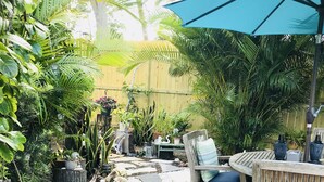 Een terras met een houten bankje, een blauwe parasol en verschillende planten in potten.