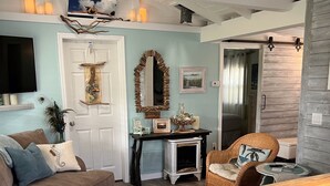 Een gezellige woonkamer met een bank, een bijzettafeltje en een rieten fauteuil. Er ligt een houten vloer en het witte plafond heeft zichtbare balken.