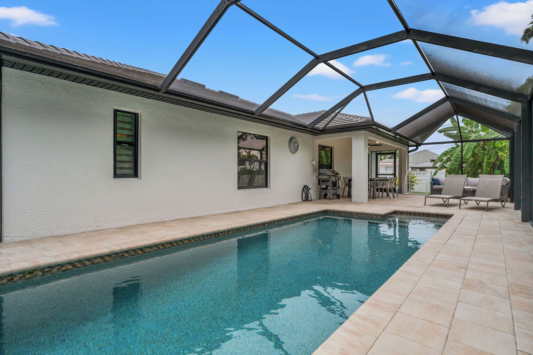 A swimming pool with a covered patio area, a clock on the wall, and outdoor furniture.