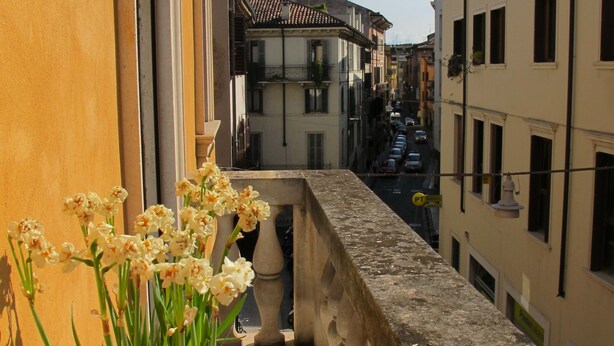 A narrow street with buildings on both sides, a balcony with flowers, and a clear sky.