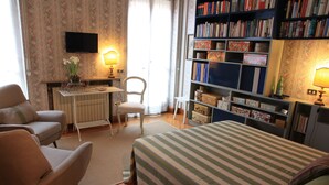A bedroom with a bed, two chairs, a desk, a bookshelf, and a TV.