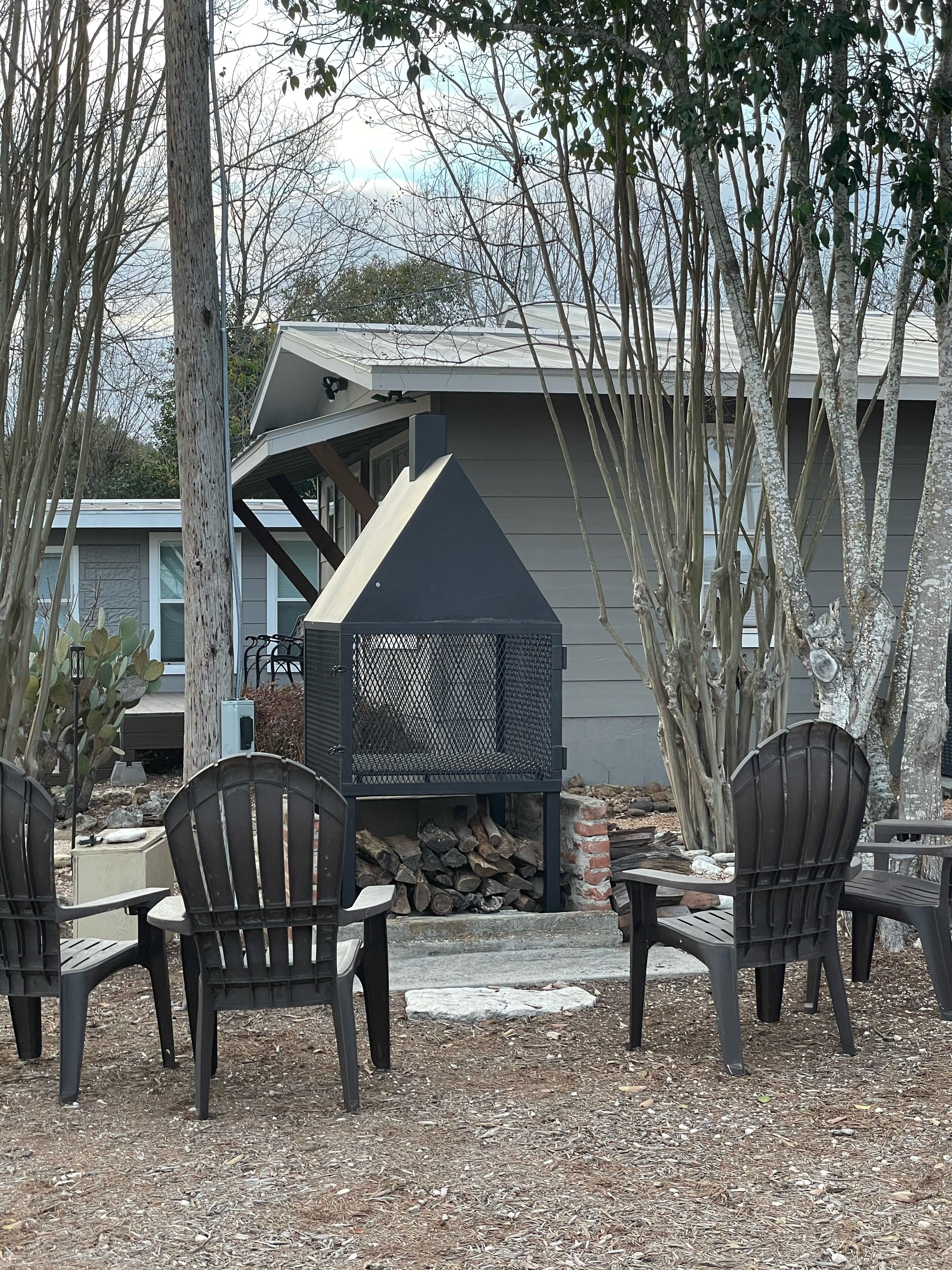 Un espace extérieur aménagé avec des chaises Adirondack, un barbecue en métal et une pile de bois de chauffage.