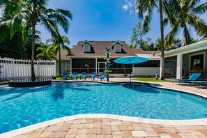 A swimming pool with a white fence, blue chairs, and a covered area with a blue umbrella.