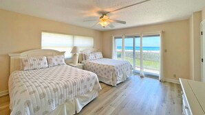 A bedroom with two beds, a ceiling fan, and a large window with a view of the beach.