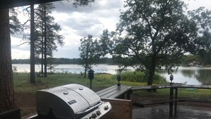 Une terrasse en bois avec un barbecue et une table, d’où l’on a vue sur un lac bordé d’arbres et sur une maison située au loin.