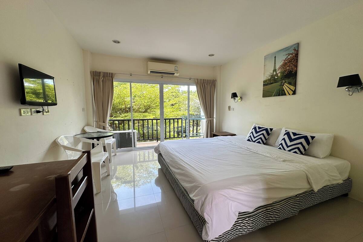 A hotel room with a bed, a desk, a chair, a TV, and a balcony with a view of trees.