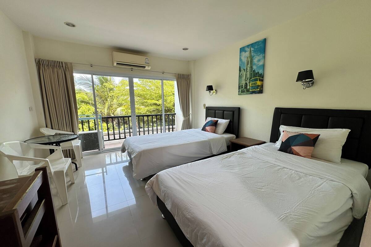 A hotel room with two beds, a small table, a chair, and a balcony with a view of greenery.