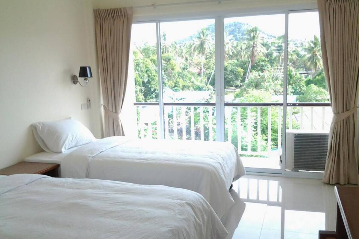 A room with two beds, a nightstand, a wall-mounted lamp, and a large window with curtains.