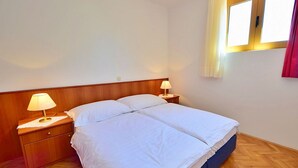 Un lit double avec du linge de lit blanc, dans une chambre au plancher en bois et dotée d’une tête de lit en bois. Il y a deux tables de chevet avec des lampes.