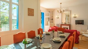 A dining area with a long table set for four, surrounded by orange chairs. There is a chandelier above the table, and a television is visible in the background. - Beautiful private villa for 10 guests with WIFI, TV and terrace (Saint-palais-sur-mer)