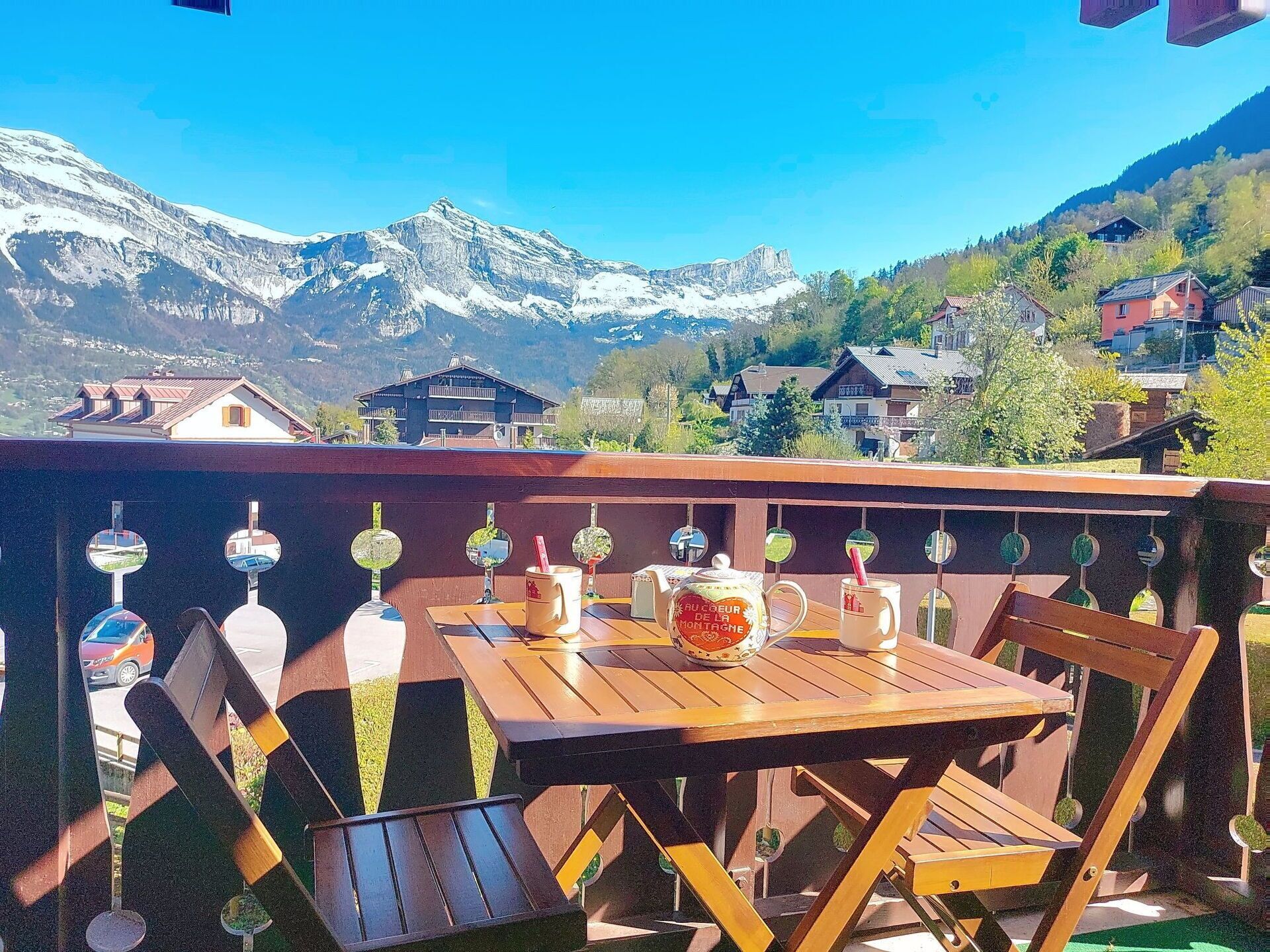 A wooden table and chairs on a balcony with a view of snow-capped mountains and houses in the background.