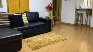 Ein Wohnzimmer mit einer schwarzen Couch, Holzboden, einem kleinen Holztisch mit Blumenvase und einem Fenster mit Blick nach draußen.