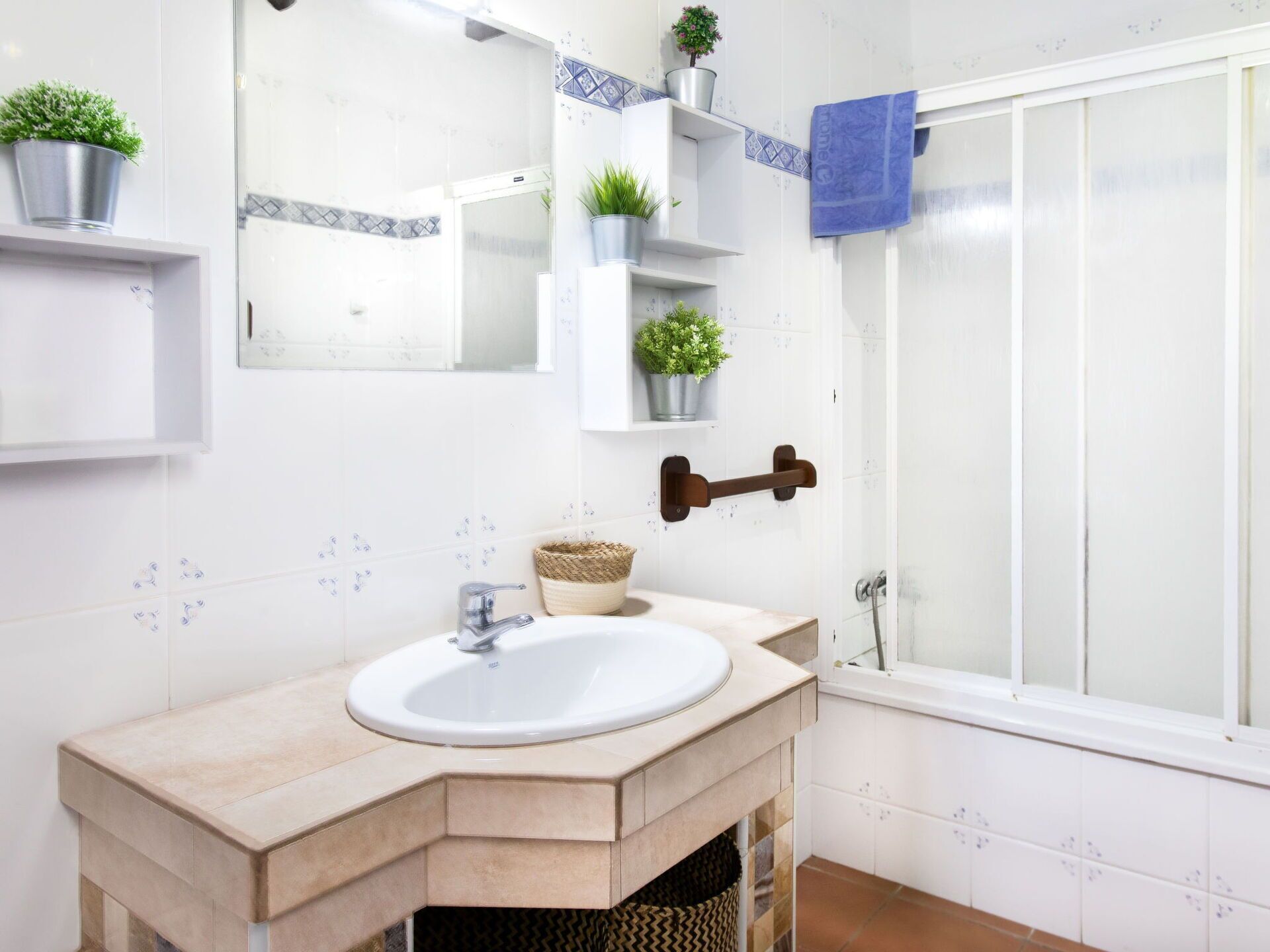 Un baño con un lavamanos blanco, un espejo y una ducha. Hay plantas en macetas sobre estantes y una toalla colgada en un perchero.