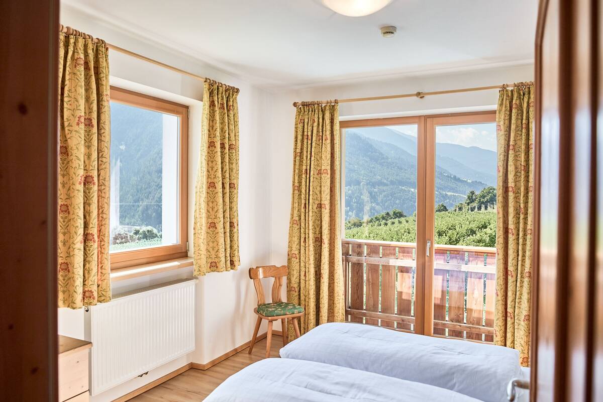 Ein Schlafzimmer mit zwei Betten, einem Holzstuhl, einem Heizkörper und einem Balkon mit Bergblick.