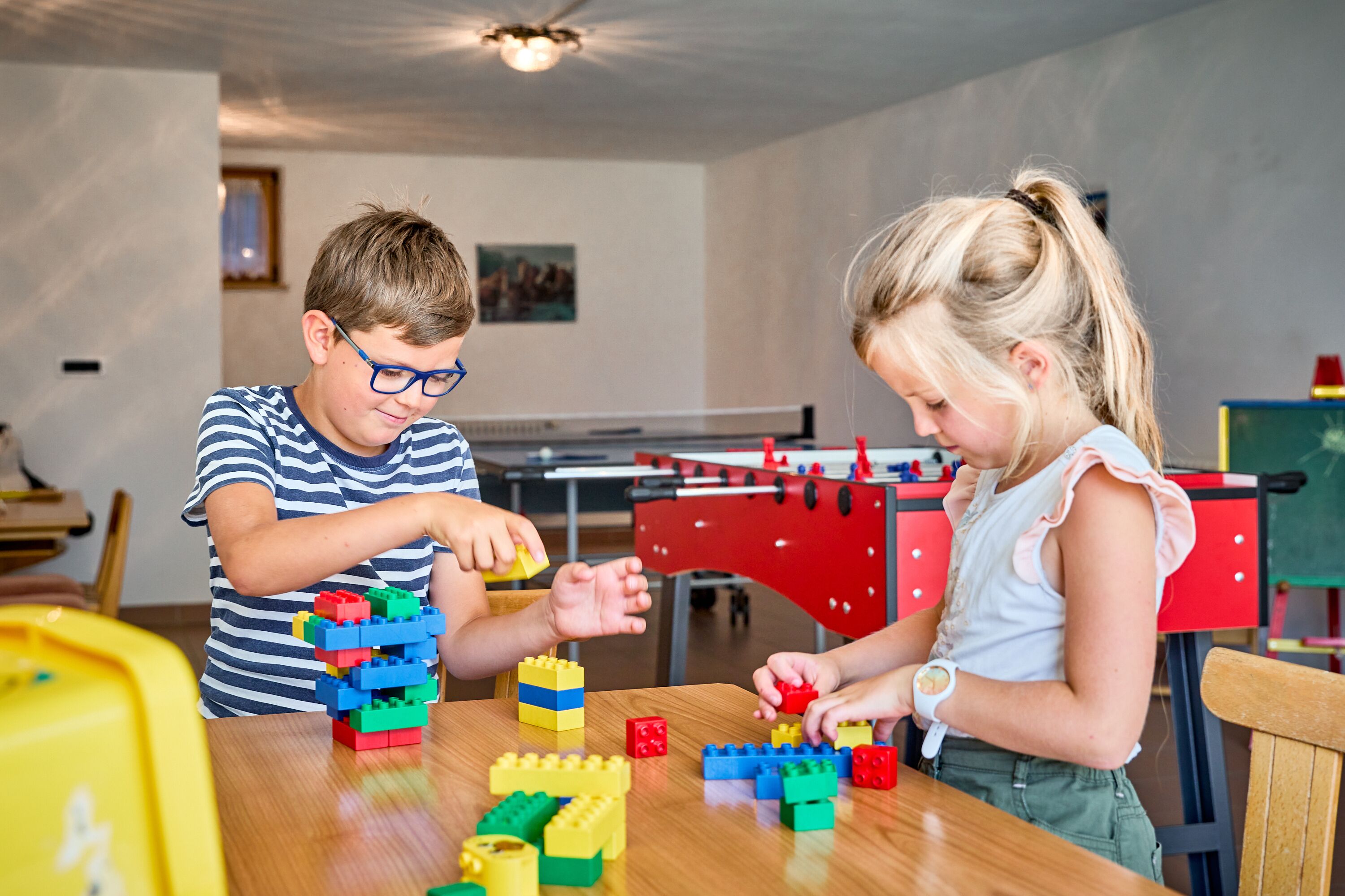 Two children playing with building blocks on a wooden table in a room with a foosball table in the background.