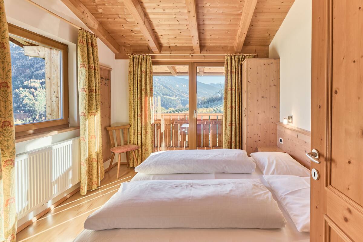 Ein Schlafzimmer mit Holzbalkendecke, einem großen Fenster mit Vorhängen, einem Bett mit weißer Bettwäsche und Bergblick.