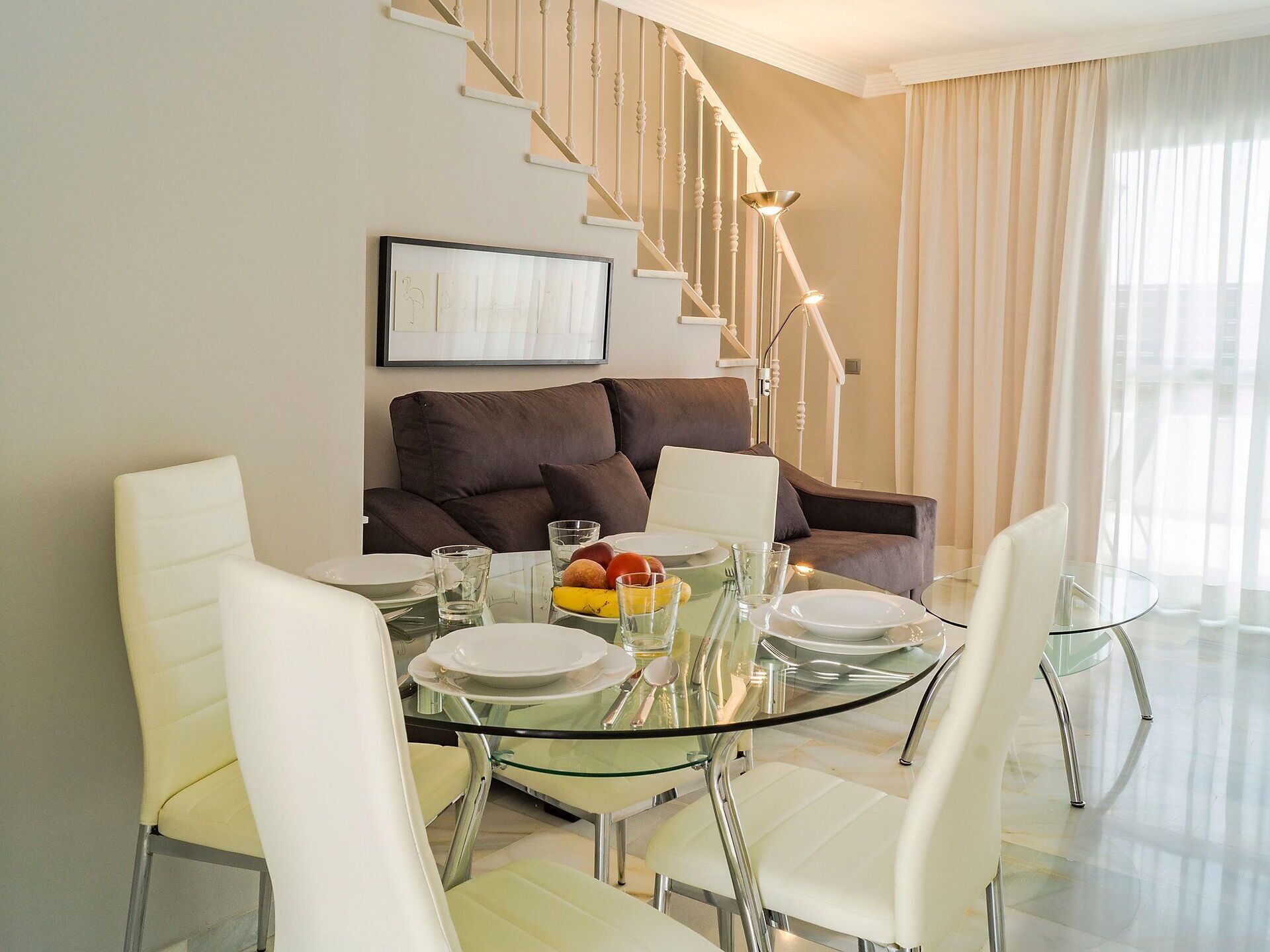 A modern dining area with a glass table, white chairs, and a brown sofa.