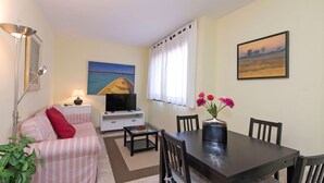 A living room with a sofa, TV, dining table, chairs, and a vase with flowers. - Nice apartment for 3 people with WIFI, A/C and TV (Barcelona)