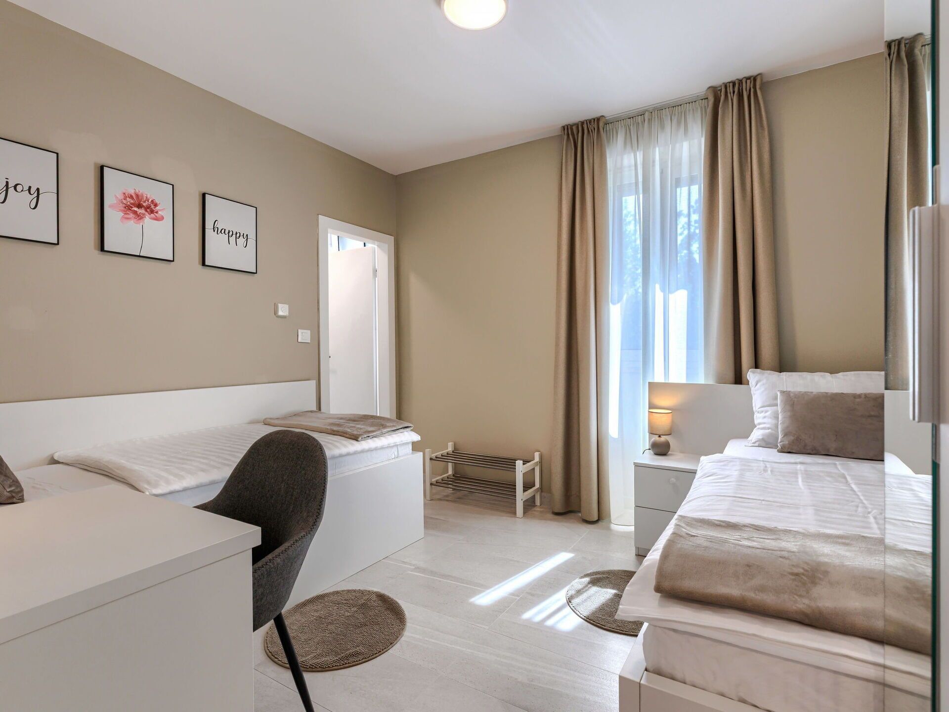 Une chambre d’hôtel moderne, équipée d’un lit, d’un bureau, d’une chaise et d’une petite table avec une lampe. Trois tableaux encadrés ornent le mur, et une fenêtre est agrémentée de rideaux.
