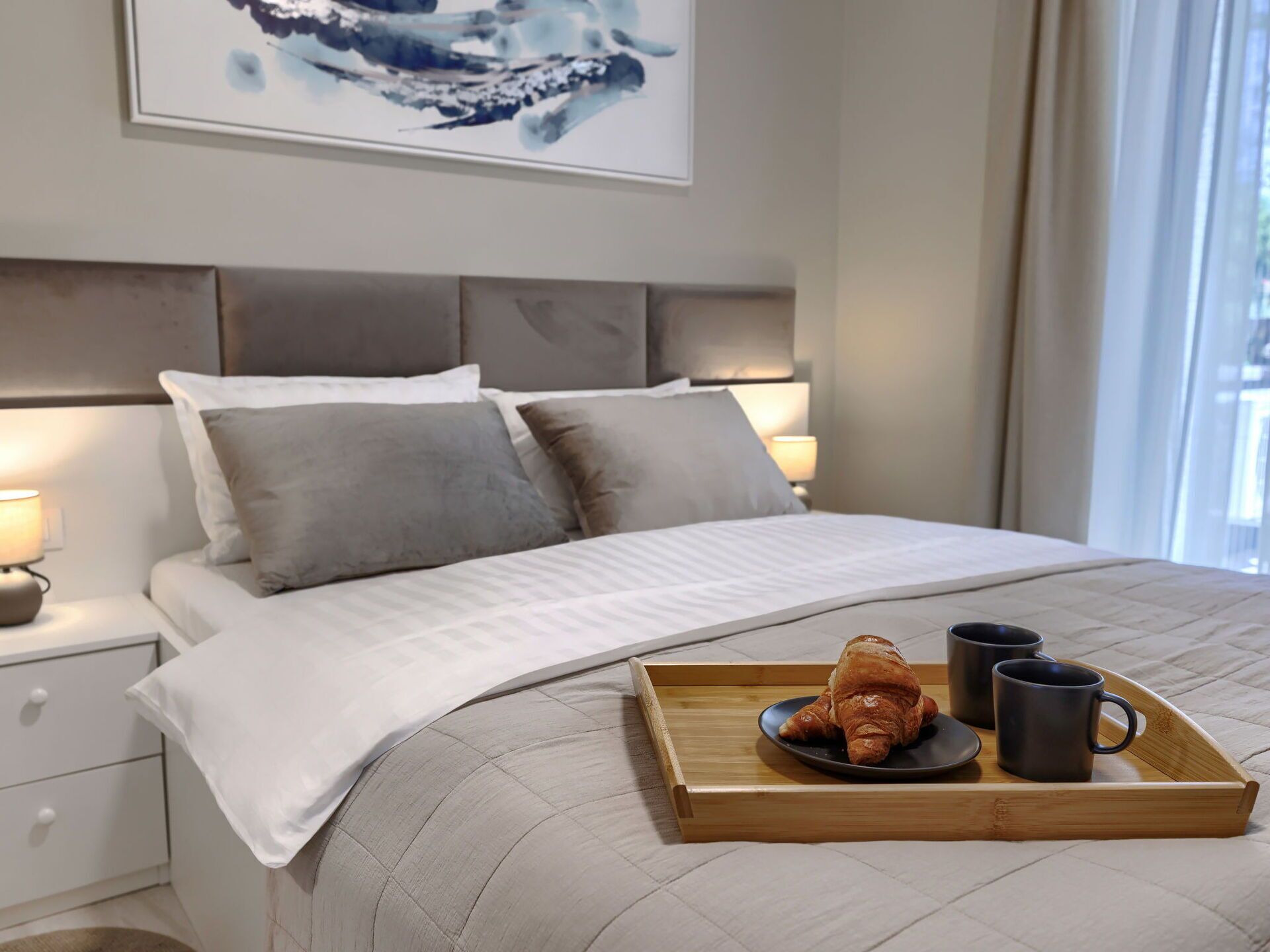 A neatly made bed with a tray of croissants and mugs, a bedside table with a lamp, and a painting on the wall.