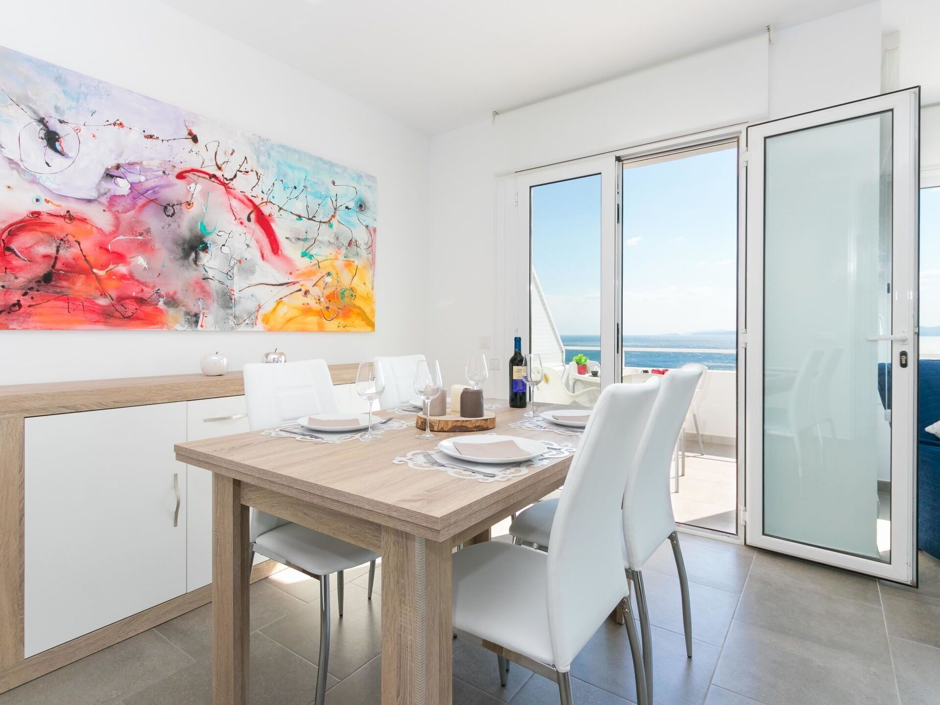 Un coin repas avec une table en bois dressée pour quatre personnes, des chaises blanches, une grande peinture abstraite accrochée au mur et une vue sur la mer par une porte vitrée.
